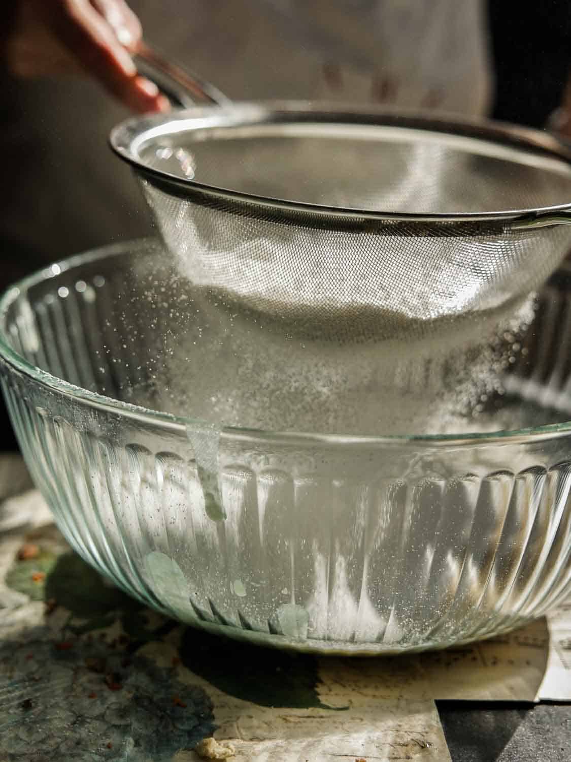 A person sifting flour through a metal sieve into a clear glass bowl. The scene is warmly lit, highlighting the falling flour and its particles. The countertop beneath appears rustic with a slight artistic touch.